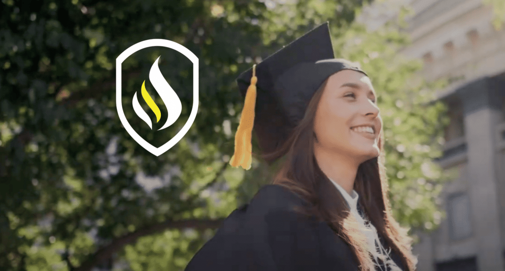 Graduate in a black cap and gown with a yellow tassel smiling outside in the sunshine with green trees.